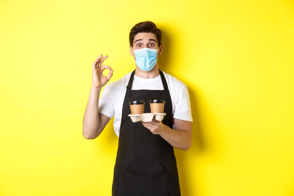 concept covid 19 cafe social distancing barista medical mask black apron guarantee safety holding takeaway cups coffee showing ok sign yellow background